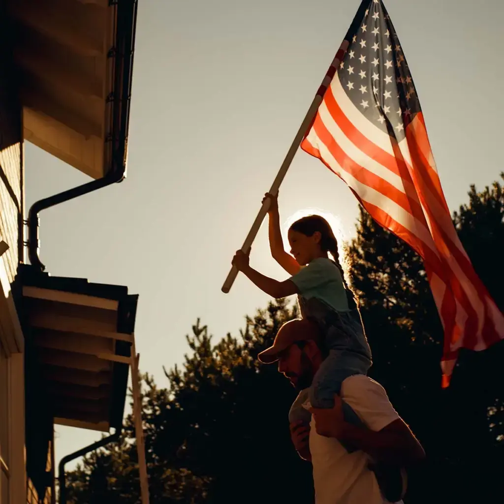 A man and a child stand side by side, joyfully holding an American flag against a clear blue sky.